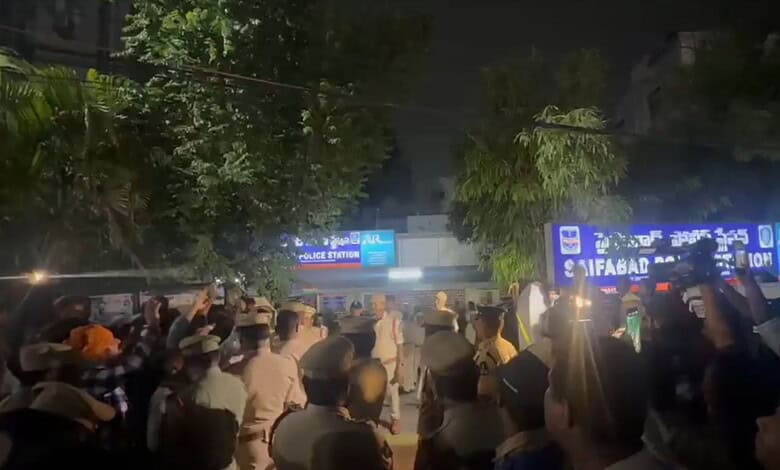 Crowd gathered outside a police station at night with illuminated signs and trees in the background.