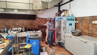 Man inspecting a refrigerator in a commercial kitchen with kitchenware and utensils around.