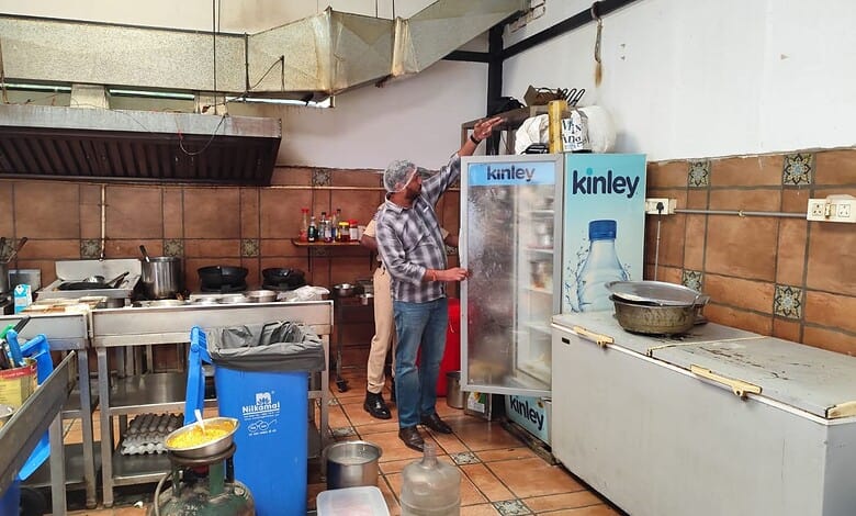 Man inspecting a refrigerator in a commercial kitchen with kitchenware and utensils around.