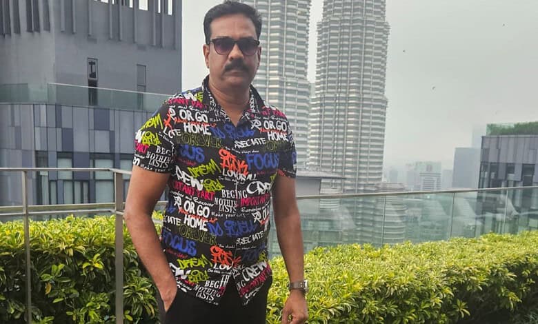 Man in colourful shirt and sunglasses standing on rooftop with cityscape background.