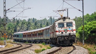 Electric train moving on railway tracks with overhead wires and green trees in background.