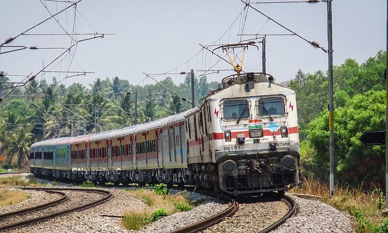 Electric train moving on railway tracks with overhead wires and green trees in background.