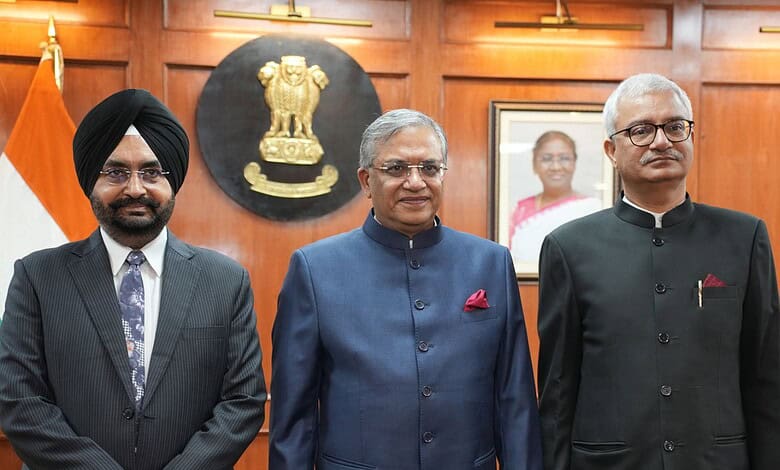 Indian election commissioners in a formal meeting room with national emblem and photo in background.