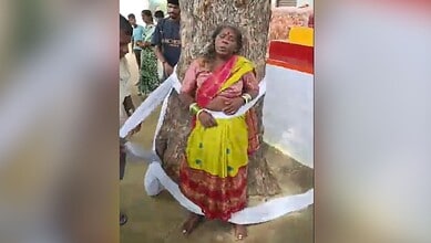 Woman in yellow and red traditional attire leaning against a tree during a cultural event.