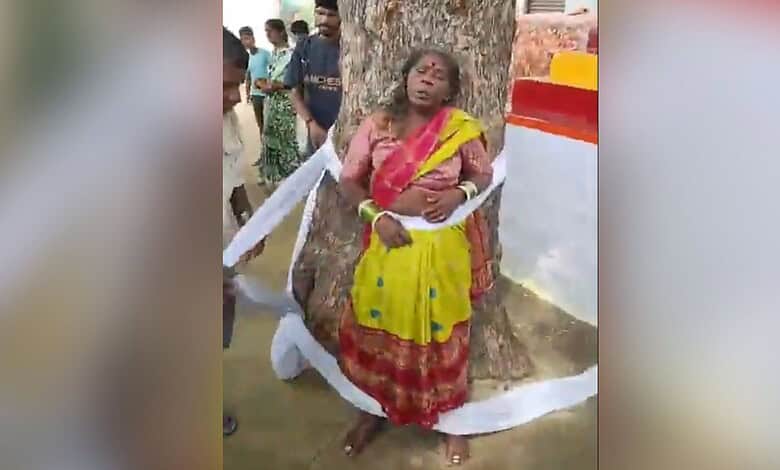 Woman in yellow and red traditional attire leaning against a tree during a cultural event.