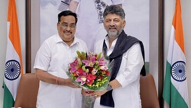 Two men exchanging a bouquet of pink lilies and greenery in an office setting with Indian flags.