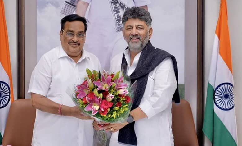 Two men exchanging a bouquet of pink lilies and greenery in an office setting with Indian flags.