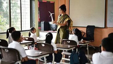Teacher instructing students in a classroom with large windows and educational posters.