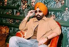 Ammy Virk wearing a turban and sunglasses, sitting on a red chair in a decorated room.