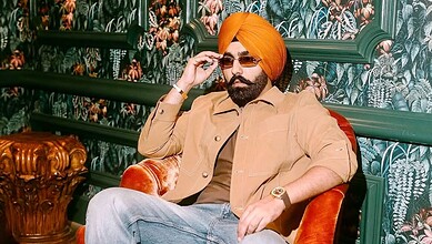 Ammy Virk wearing a turban and sunglasses, sitting on a red chair in a decorated room.