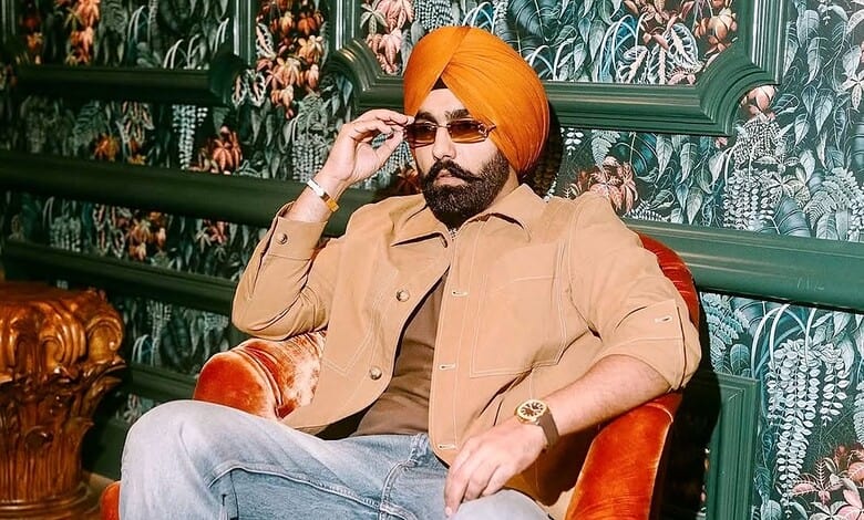 Ammy Virk wearing a turban and sunglasses, sitting on a red chair in a decorated room.