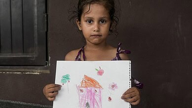 Young girl holding a colourful drawing of a house and flowers, looking serious.