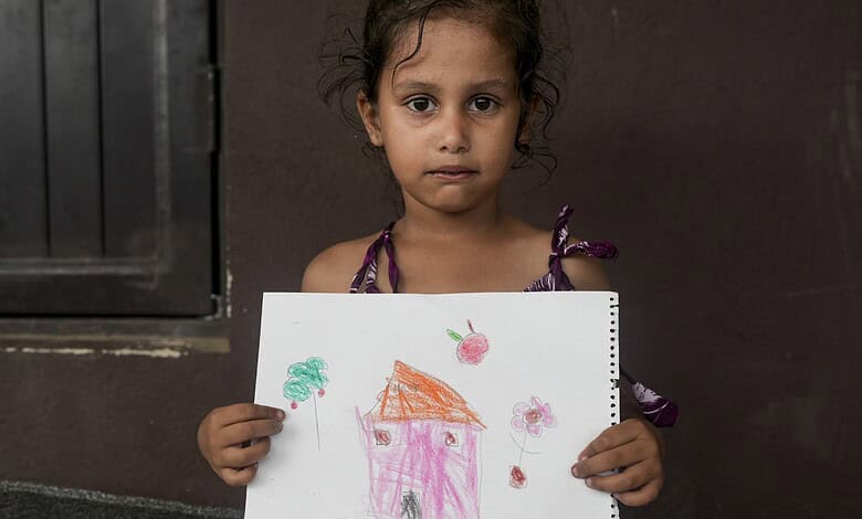 Young girl holding a colourful drawing of a house and flowers, looking serious.