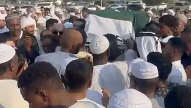 Large crowd of people attending a funeral procession outdoors in a sunny setting.