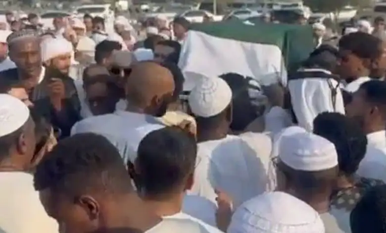 Large crowd of people attending a funeral procession outdoors in a sunny setting.