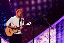 Ed Sheeran playing guitar and singing on stage during a concert.