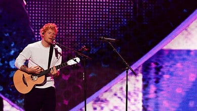Ed Sheeran playing guitar and singing on stage during a concert.