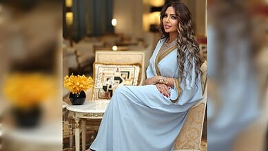 Beautiful woman with long wavy hair wearing a light blue traditional dress, sitting in a luxurious room.