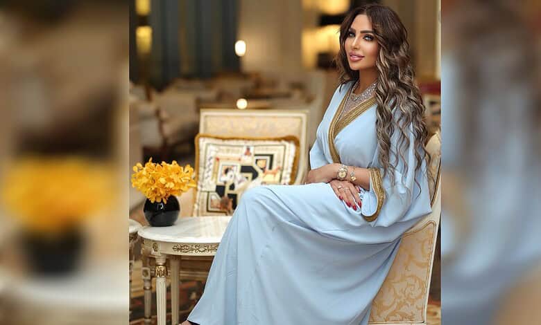 Beautiful woman with long wavy hair wearing a light blue traditional dress, sitting in a luxurious room.