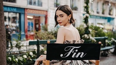 Lily Collins with dark hair and elegant makeup, sitting outdoors with a 'Fin' sign.