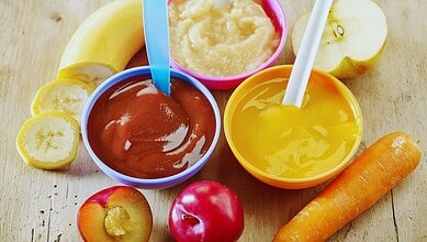 Three bowls of homemade baby food purees (plum, apple, and carrot) surrounded by fresh banana, apple, plums, and a carrot on a wooden table.