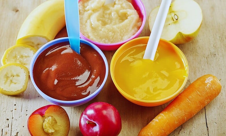 Three bowls of homemade baby food purees (plum, apple, and carrot) surrounded by fresh banana, apple, plums, and a carrot on a wooden table.