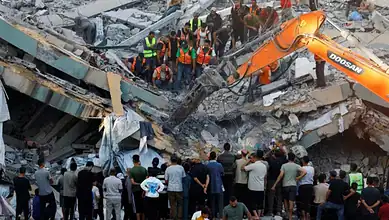 Rescue workers and onlookers at the site of a building collapse in Gaza, with heavy machinery and debris.