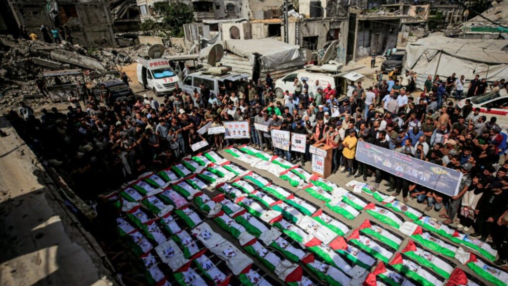 Bodies wrapped in Palestinian flags laid out during a mass funeral in Gaza City’s Al-Zaytoun neighbourhood, surrounded by mourners and war-damaged buildings.