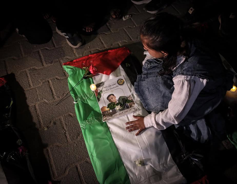 A young girl mourns beside a shrouded body draped in a Palestinian flag, with a photograph and flowers placed on top.