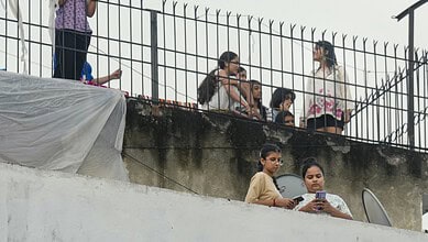 Group of young women and girls on a rooftop, some using mobile phones, with a metal fence in the scene.