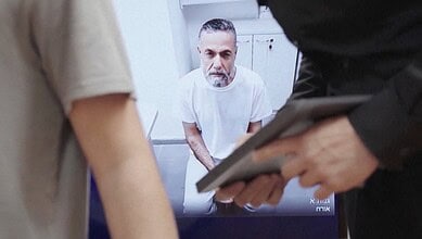 Gaza doctor giving testimony in prison, surrounded by officials, holding a document.
