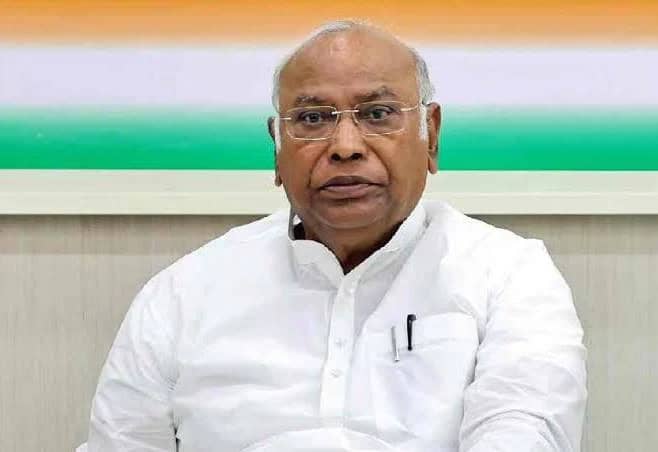 Indian politician wearing white shirt and glasses, sitting indoors with a neutral expression.