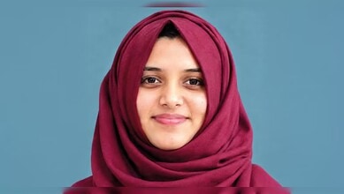 Close-up of a young woman wearing a maroon hijab, smiling softly against a blue background.