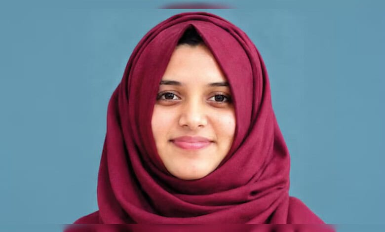 Close-up of a young woman wearing a maroon hijab, smiling softly against a blue background.