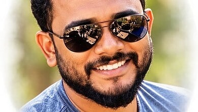 Young man with a beard and sunglasses smiling outdoors in sunlight.