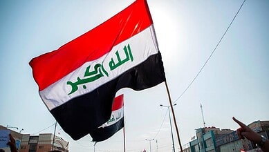 Iraqi flag waving in the sky during daytime with cityscape background.