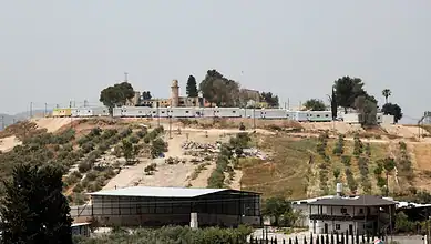 A fortified area with walls and watchtowers on a hilltop, surrounded by trees and buildings.