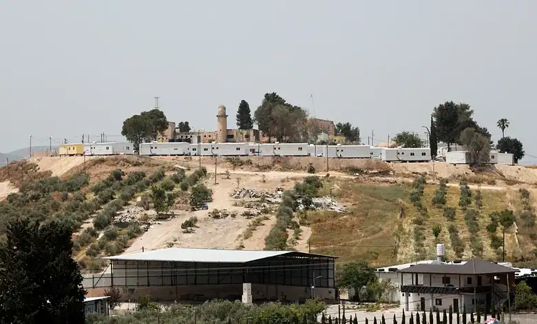A fortified area with walls and watchtowers on a hilltop, surrounded by trees and buildings.