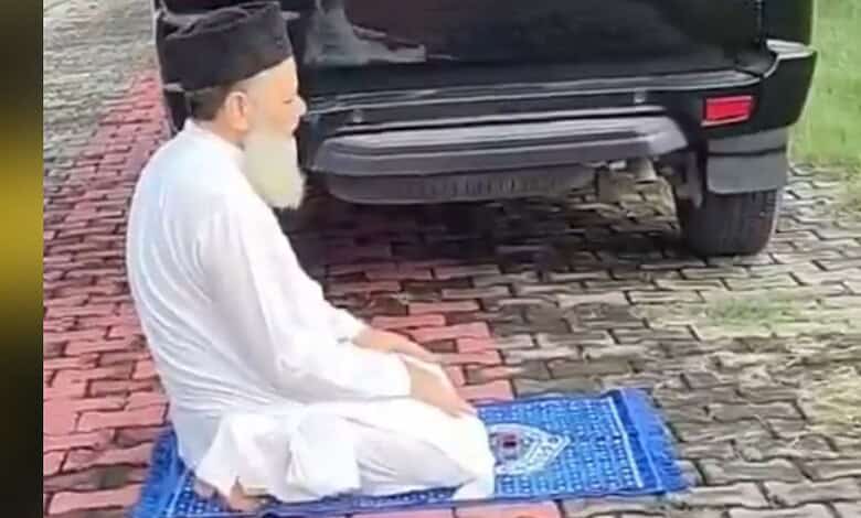 Man in white traditional attire offering prayer on a blue prayer mat outdoors near a black vehicle.