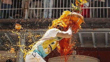 Woman participating in Dahi Handi festival, breaking pot with a crowd in background.
