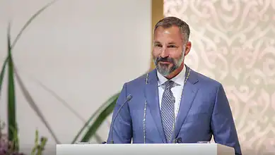 Mawlana Hazar Imam speaking at a formal event in a blue suit with a beard and smiling.