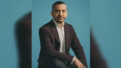Mehdi Hasan wearing a dark blazer and white shirt against a blue background.
