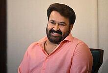 Mohanlal smiling in a pink shirt, seated indoors, promoting the Dubai entertainment event.