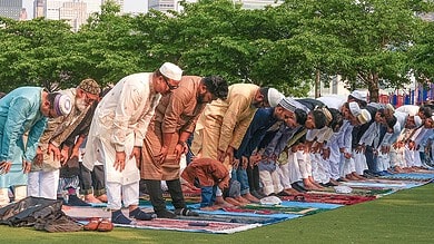 Muslims performing Eid prayer outdoors on a green field with city buildings in the background.