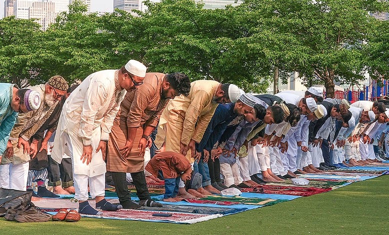 Muslims performing Eid prayer outdoors on a green field with city buildings in the background.