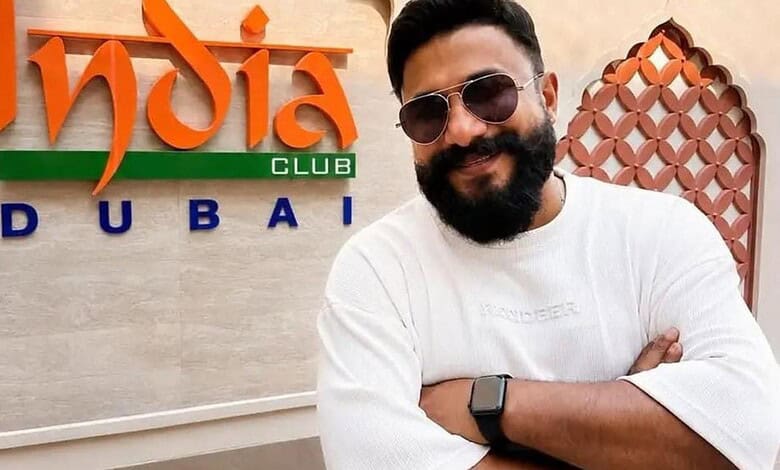 Indian singer with beard and sunglasses smiling in Dubai, in front of India Club Dubai sign.