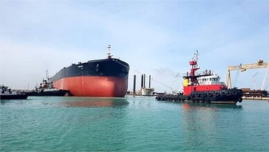 Large cargo ship in Persian Gulf with tugboats and port infrastructure visible.