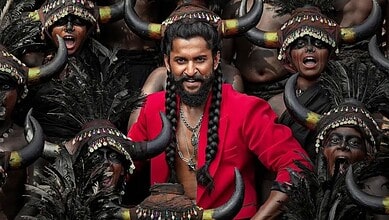 Man in red shirt performing tribal dance with dancers wearing horns and traditional attire.