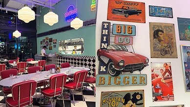Retro-themed diner interior with vintage car and 1960s posters, vibrant neon lights, and red chairs.