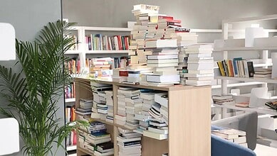 Overflowing bookshelves in a library with a large stack of books on a table.